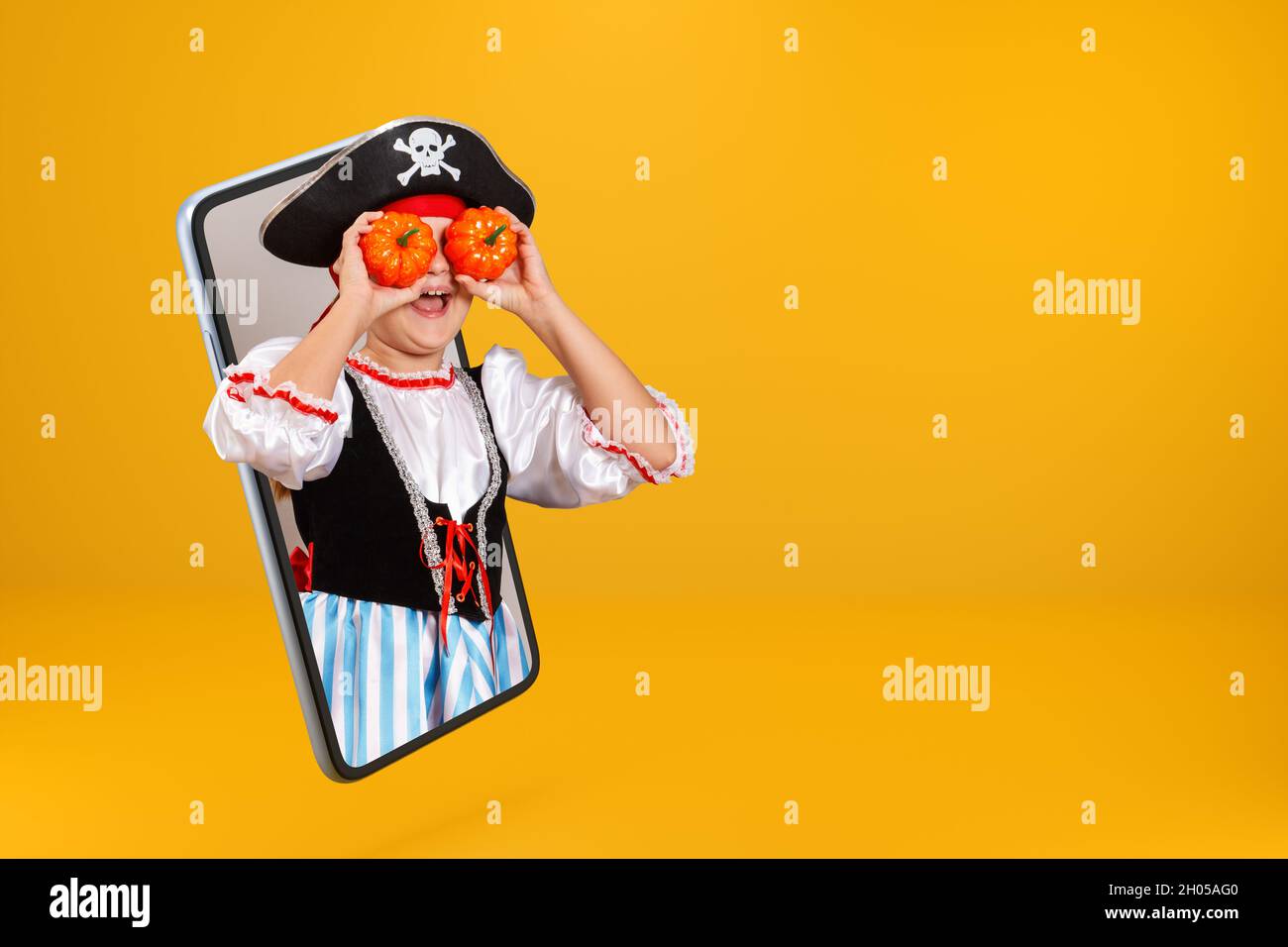 Ein Kind im Karnevalskostüm eines Piraten schaut von einem Smartphone aus. Kleines Mädchen an Halloween online auf einem Handy-Bildschirm und hält Kürbisse inste Stockfoto