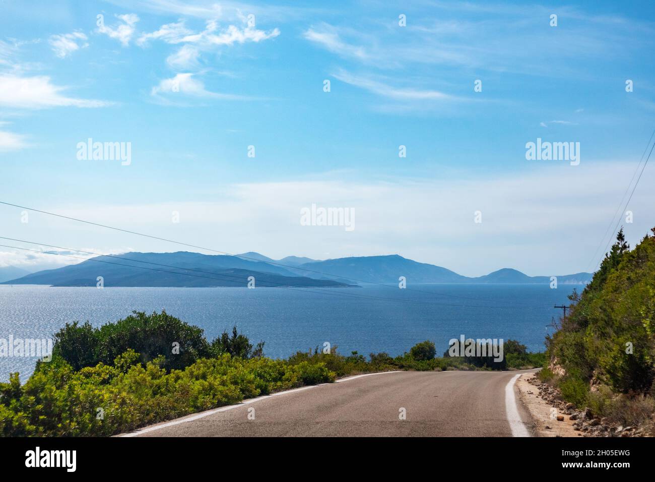 Sie fahren auf einer hohen, steilen Straße mit grünen, lebhaften Hügeln, blauen, fernen Inselsilhouetten und einem klaren, blauen Himmel. Reisen Lefkada Island, Griechenland. Sonnige Sommerlandschaft Stockfoto