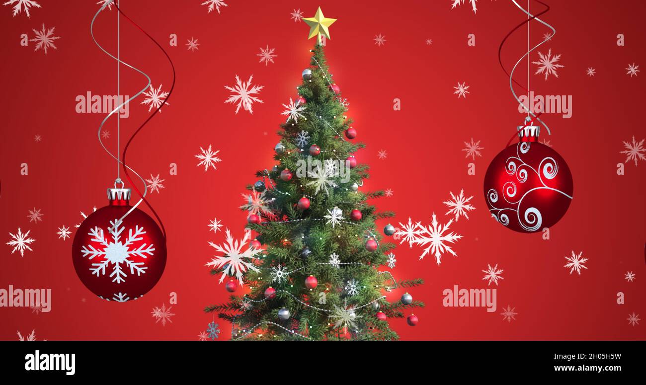 Bild von Schnee fällt über weihnachtskugeln und weihnachtsbaum auf rotem Hintergrund Stockfoto