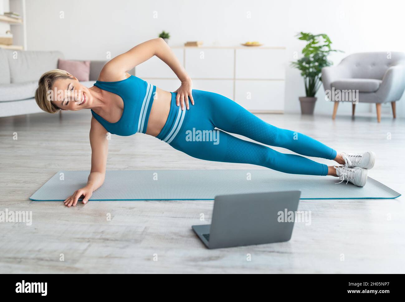 Ganzkörperlänge von Happy Fit Frau Training Stehen in Side Plank Position auf dem Boden Yoga-Matte im Wohnzimmer, Blick auf Computer-Bildschirm beobachten T Stockfoto