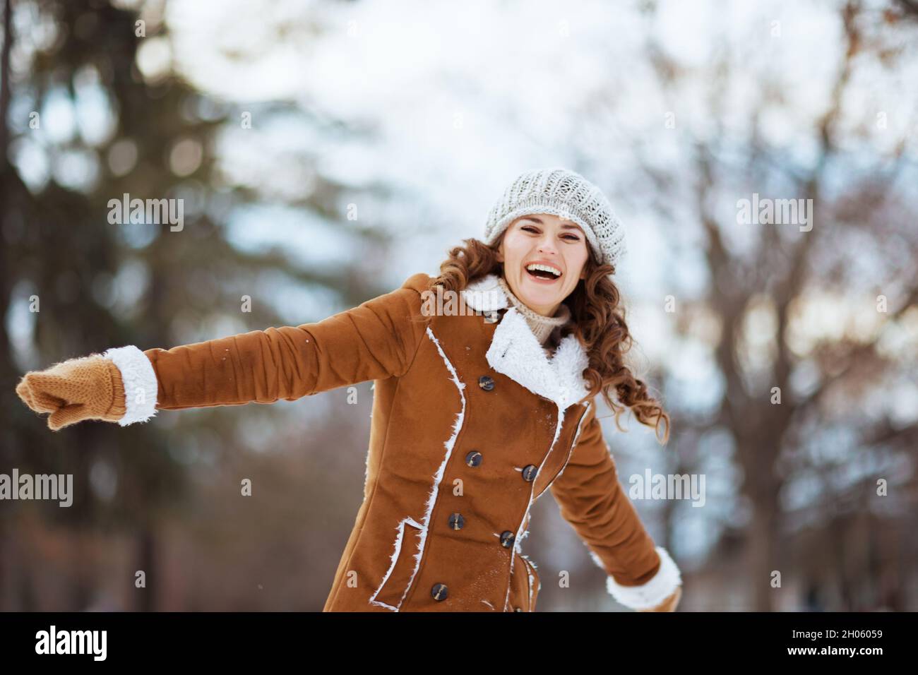 Happy stylish Frau mit Fäustlingen in einem gestrickten Hut und Schaffell Mantel frohlocken im Freien im Stadtpark im Winter. Stockfoto