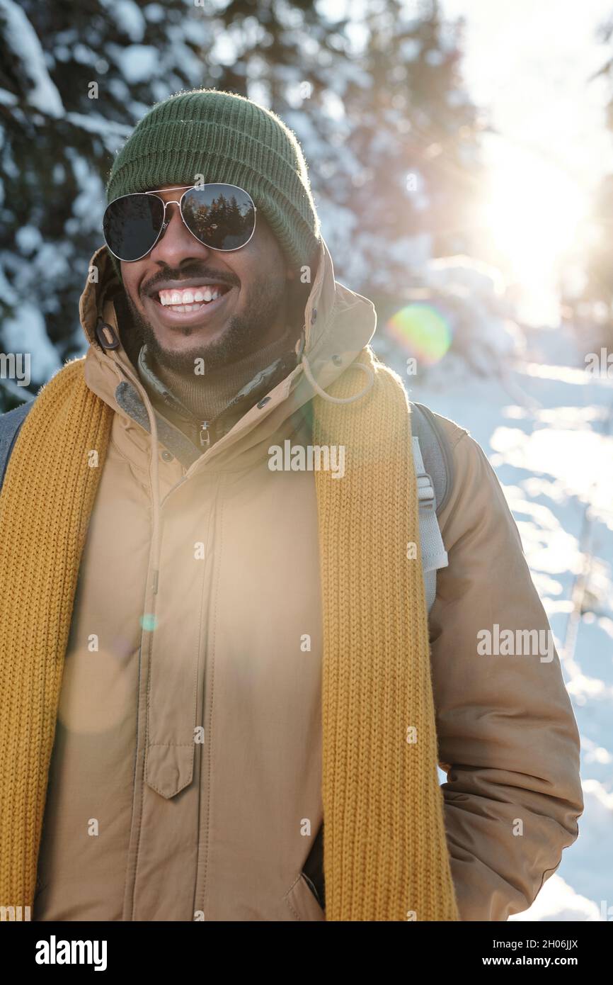 Glücklicher junger Afrikaner in Winterkleidung und Sonnenbrillen, die im Winterwald chillen Stockfoto