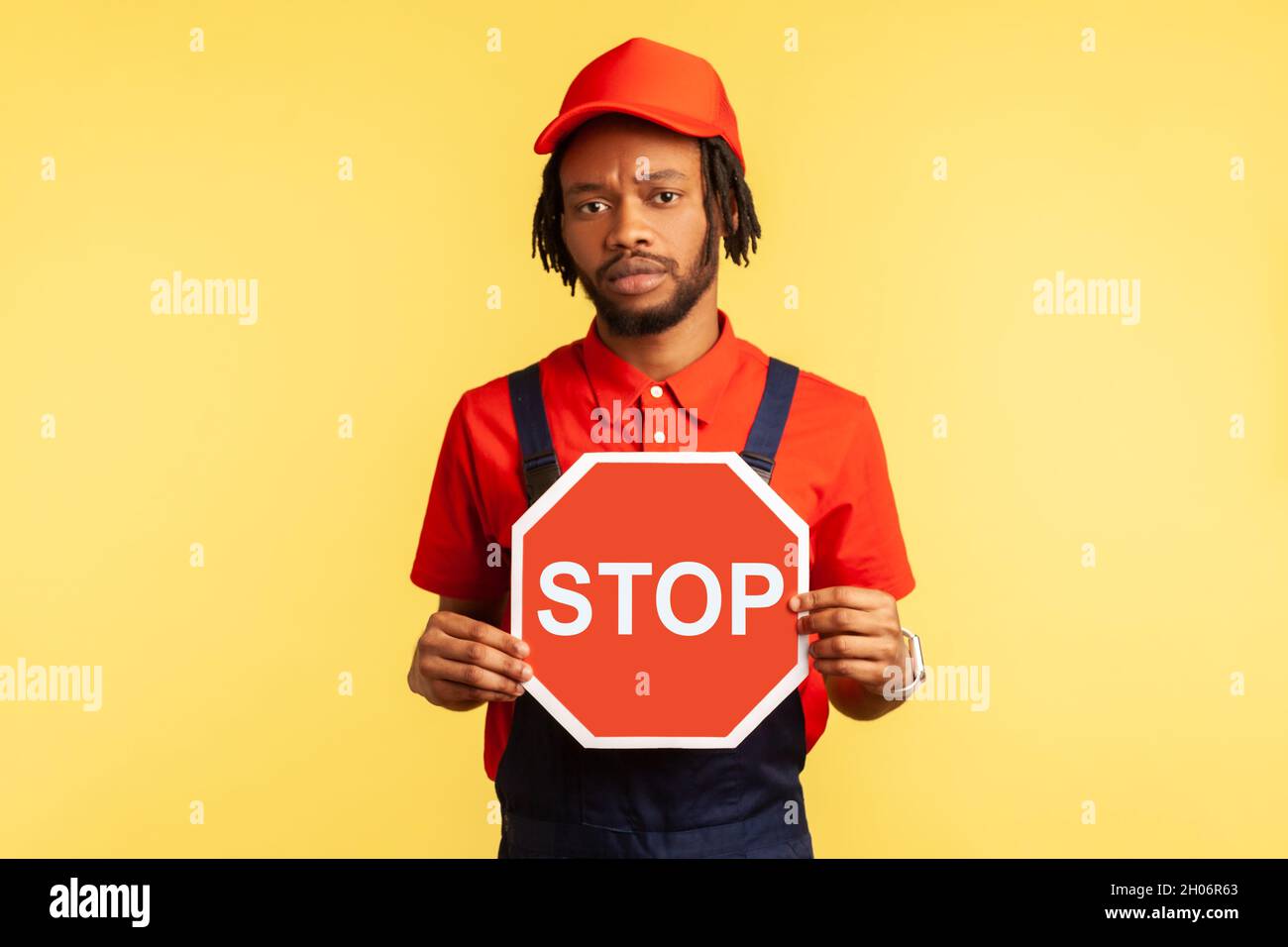 Porträt eines Handwerkers mit blauen Overalls, rotem Hut und T-Shirt mit rotem Stoppschild, Verbote, ernsthafter Blick auf die Kamera. Innenaufnahme des Studios isoliert auf gelbem Hintergrund. Stockfoto