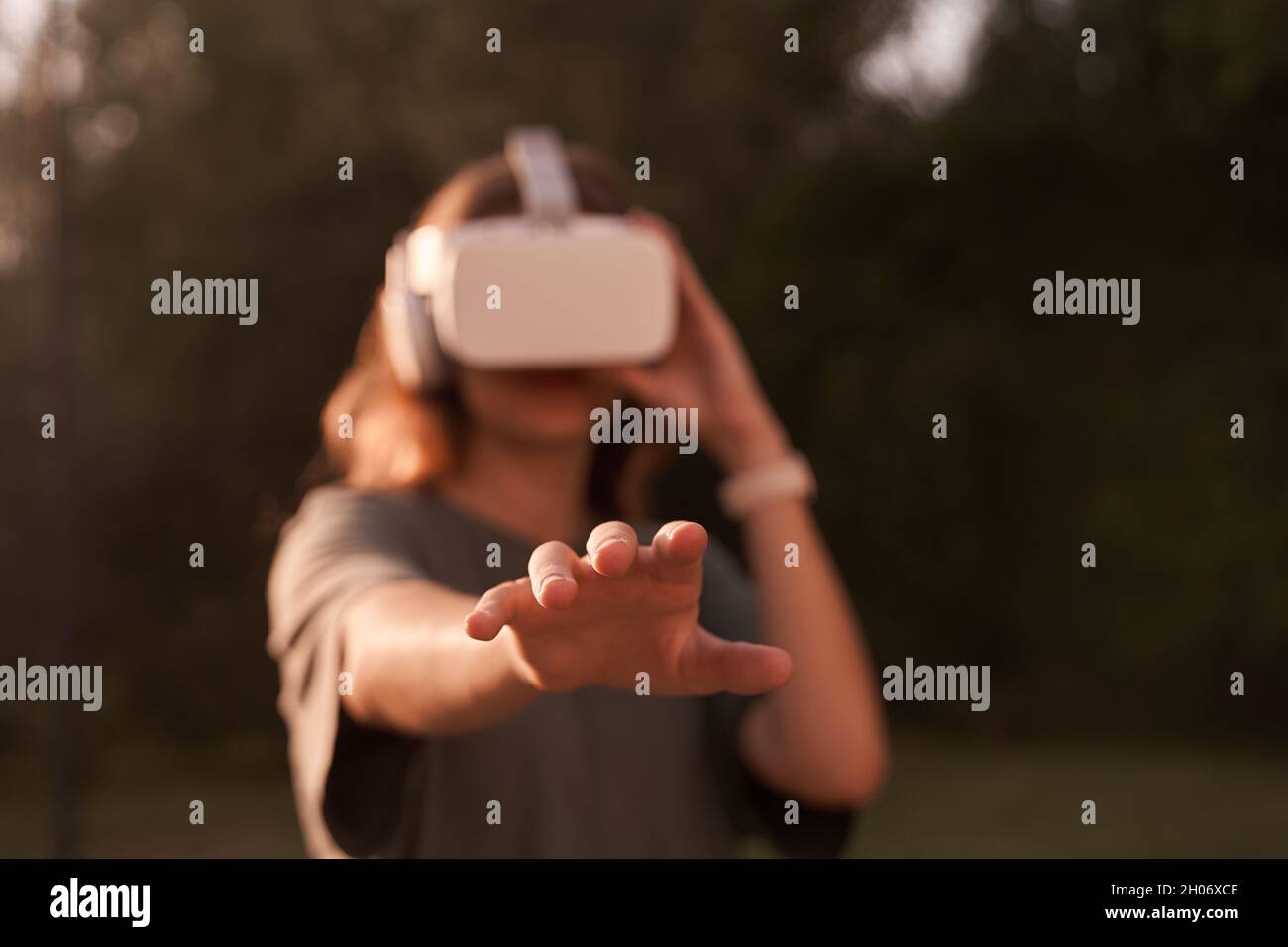 Nahaufnahme eines Mädchens, das mit den Händen eine Virtual-Reality-Brille auf dem Kopf hält und auf einem Hintergrund von grünen Bäumen steht. Er reicht eine Hand an die Kamera. Hochwertige Fotos Stockfoto