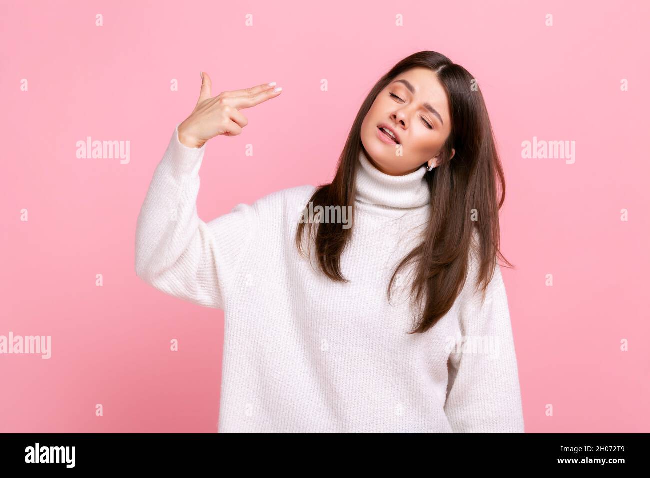 Unglückliche Frau steht mit der Fingerpistole auf den Kopf gerichtet, gelangweilt oder müde, schwer mit dem Problem umzugehen, trägt weißen Pullover im lässigen Stil. Innenaufnahme des Studios isoliert auf rosa Hintergrund. Stockfoto