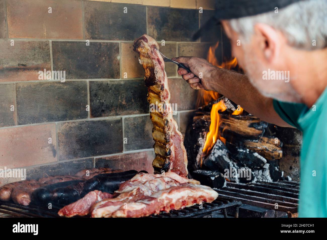 Asado. Argentinischer reifer Mann, der auf dem Grill seines Hauses gegrillt wird. Er prodding das Fleisch mit einer Gabel. Stockfoto