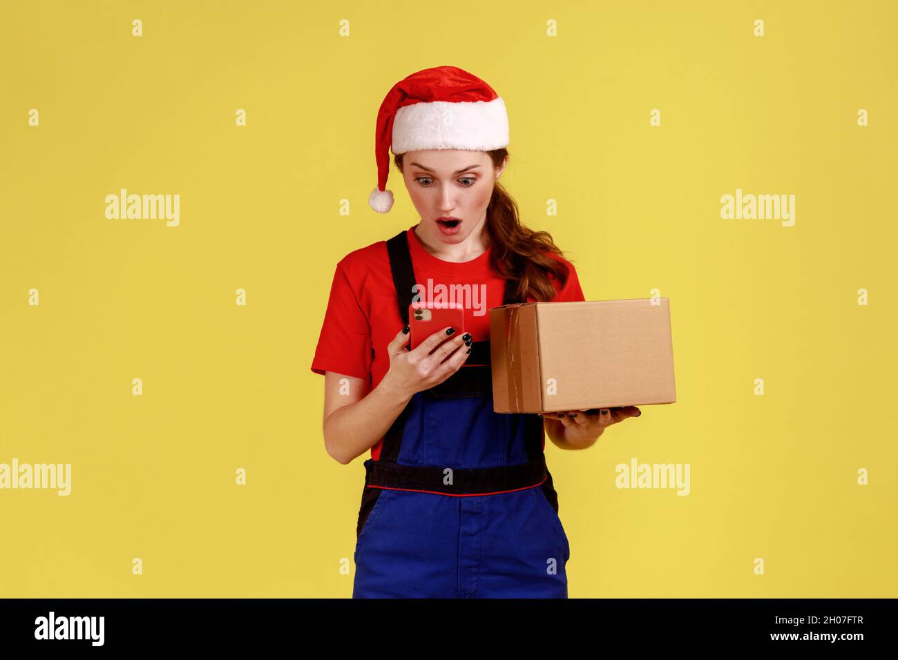 Überraschte die Zustellerin mit dem Paket in den Händen, benutzte das Handy, hatte einen Fehler mit der Kundenadresse, trug blaue Overalls und einen weihnachtsmann-Hut. Innenaufnahme des Studios isoliert auf gelbem Hintergrund. Stockfoto