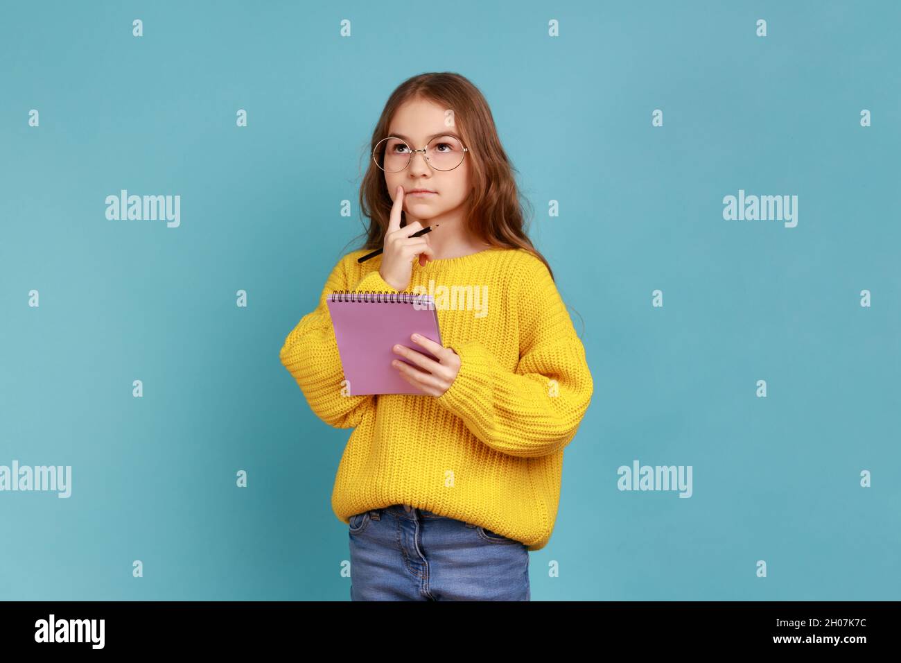 Porträt eines kleinen Mädchens, das im Notizbuch schreibt, denkt, Notizen macht, Schulkind Hausaufgaben macht, gelben Pullover im lässigen Stil trägt. Innenaufnahme des Studios isoliert auf blauem Hintergrund. Stockfoto