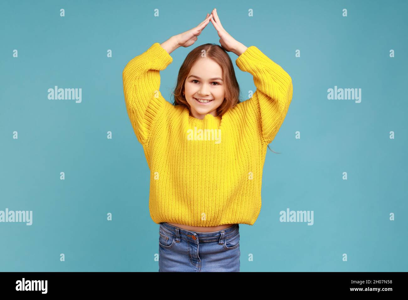 Kleines Mädchen tut Hausdach Geste mit den Händen über den Kopf und lachen, Kinderbetreuung und Schutz, tragen gelben lässigen Stil Pullover. Innenaufnahme des Studios isoliert auf blauem Hintergrund. Stockfoto