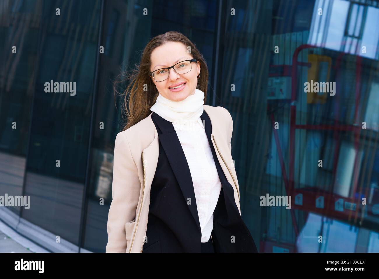 Porträt einer glücklichen erfolgreichen kaukasischen Ethnie Frau in Brille, steht in Mantel und Anzug vor der Kulisse eines Bürogebäudes an einem sonnigen Tag. Professioneller Anwalt und Finanzier echte Person Stockfoto