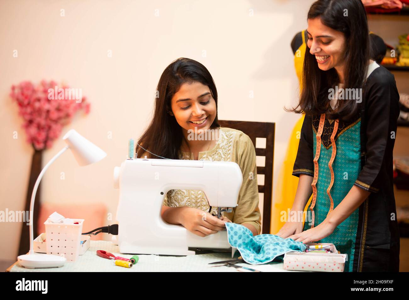 Junge indische glückliche Frauen, Schneider, Modedesignerin, mit Nähmaschine, miteinander reden und lächeln, sitzen hinter ihrem Schreibtisch in ihrem Kleidungsstück Schneiderei w Stockfoto