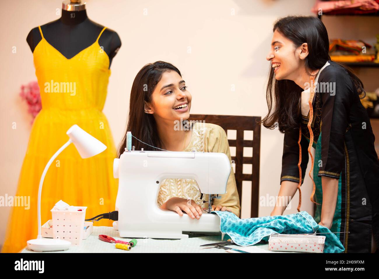 Junge indische glückliche Frauen, Schneiderin, Modedesignerin, mit Nähmaschine, miteinander reden und lachen, sitzen hinter ihrem Schreibtisch in ihrem Kleidungsstück Schneiderei Stockfoto