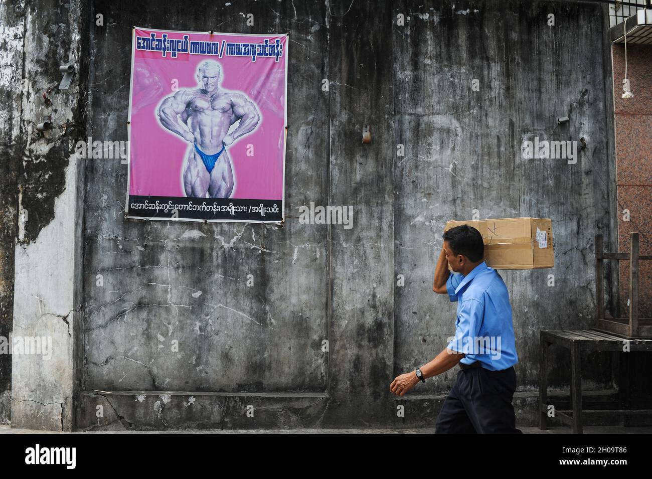 '11.10.2013, Myanmar, , Yangon - Ein Paketdienstleister trägt ein Paket auf seiner Schulter und geht mit einem Plakat, das einen Bodybuilding-Abend anwirbt, an einer Wand vorbei Stockfoto