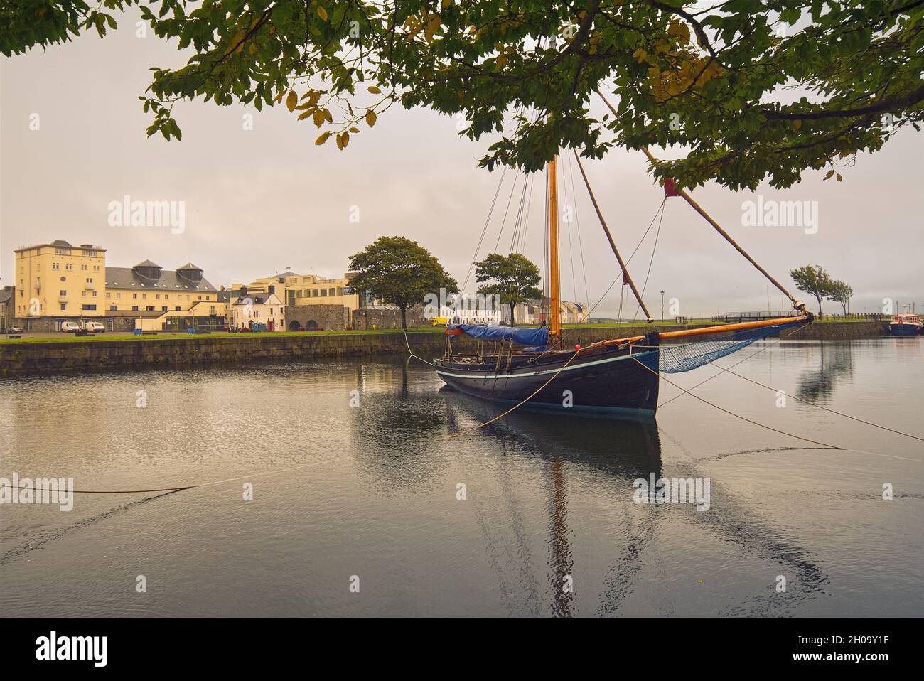 Wunderschöne Stadtlandschaft mit einem alten hölzernen Fischerboot namens Galway Hooker im Corrib River mit bunten Häusern im Hintergrund bei Claddagh in Galway City, Irland Stockfoto