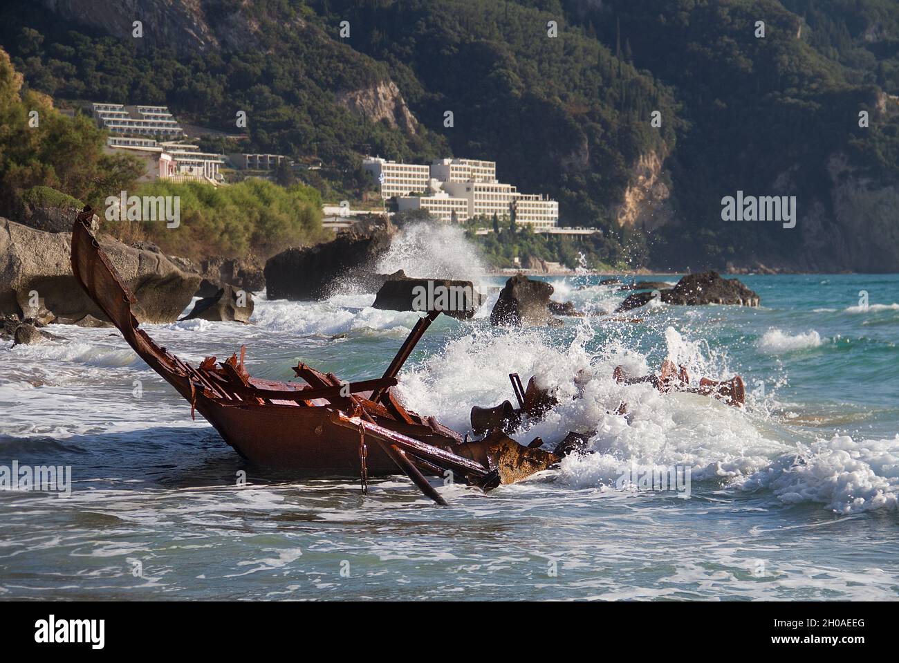 Wellen, die auf dem rostigen Wrack eines gebrochenen, gestrandeten Fischerbootes plantschen, im Hintergrund ein Touristenort Stockfoto