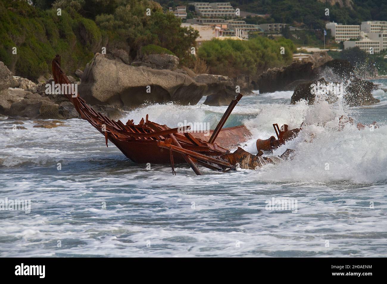 Wellen, die auf dem rostigen Wrack eines gebrochenen, gestrandeten Fischerbootes plantschen, im Hintergrund ein Touristenort Stockfoto