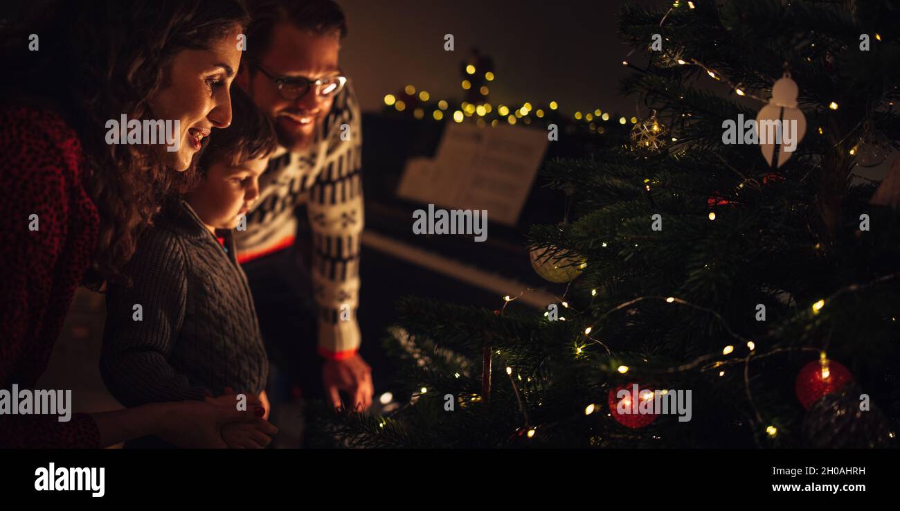 Kleine Familie, die zusammen Weihnachtsbaum schmückt. Eltern mit Kind, die zu Hause einen geschmückten Weihnachtsbaum betrachten. Stockfoto