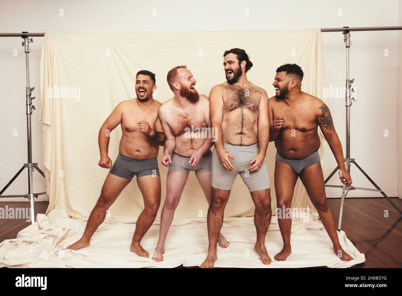 Natürlich zu sein macht Spaß. Vier körperpositive junge Männer lachen fröhlich, während sie in Unterwäsche zusammen stehen. Selbstbewusste junge Männer umarmen ihre Stockfoto