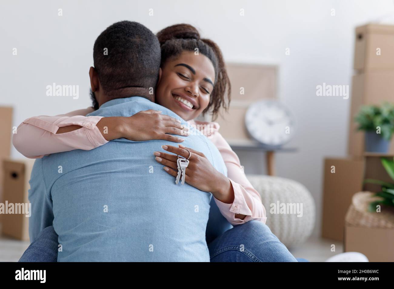 Die lächelnde, tausendjährige afroamerikanische Frau umarmte den Mann in einer neuen Wohnung und die Dame hielt die Schlüssel im Zimmer mit den Boxen Stockfoto