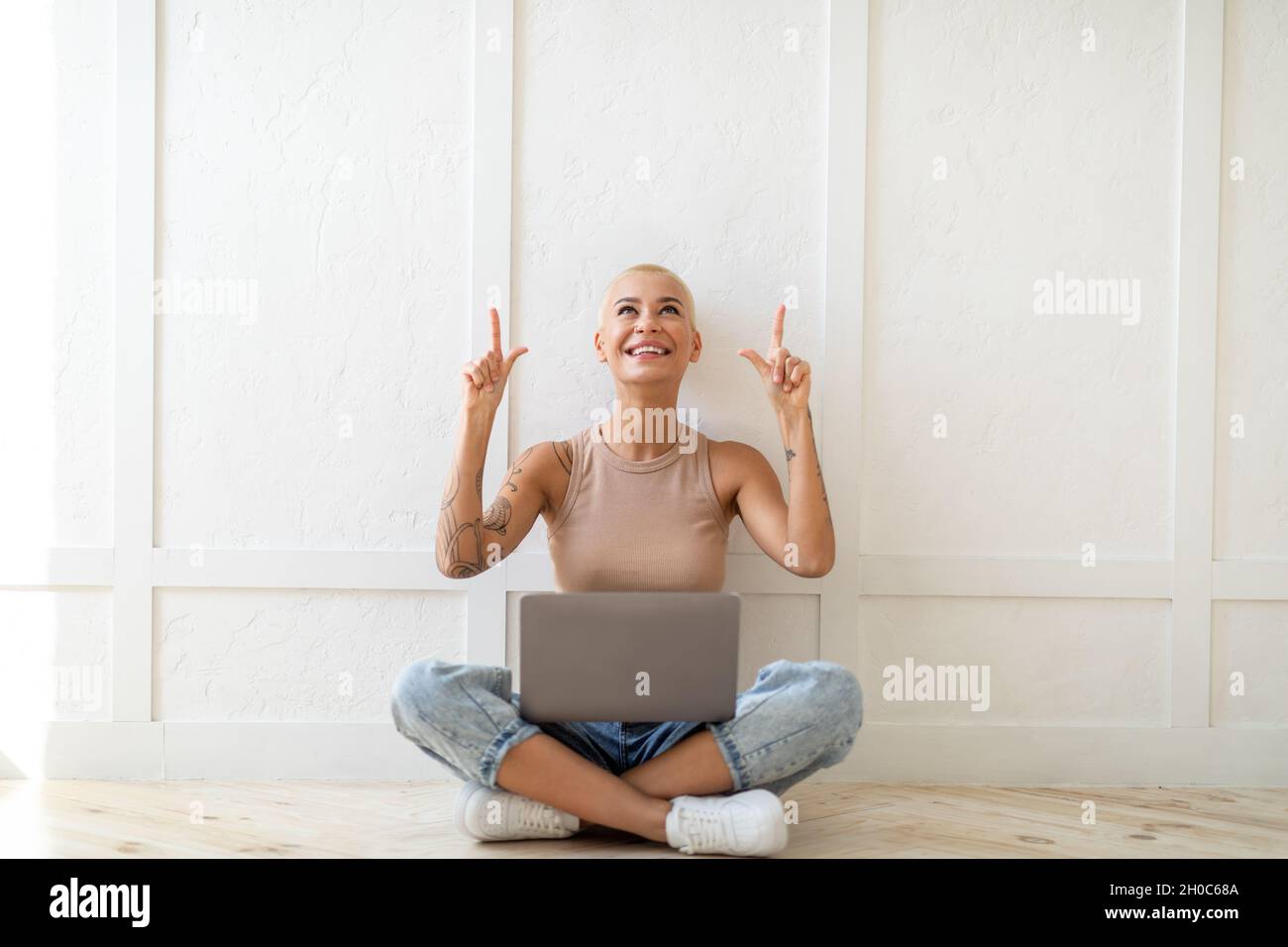 Überprüfen Sie dies. Aufgeregte junge Dame, die mit einem Laptop sitzt und auf freien Platz über ihrem Kopf über der weißen Wand zeigt Stockfoto