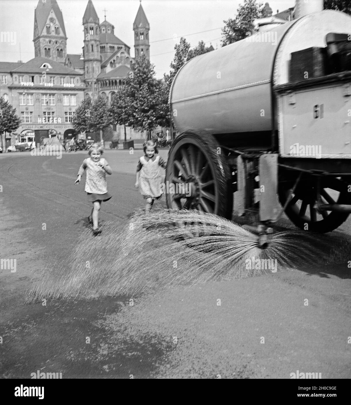 Kinder laufen dem Stadtreinigungswagen hinterher, der den zentralen Platz in Köln, den Neumarkt, naß säubert, 1930er Jahre. Kinder haben Spaß, während die öffentliche Reinigung Duschen der zentralen Platz von Köln, der Neumarkt, 1930er Jahre. Stockfoto