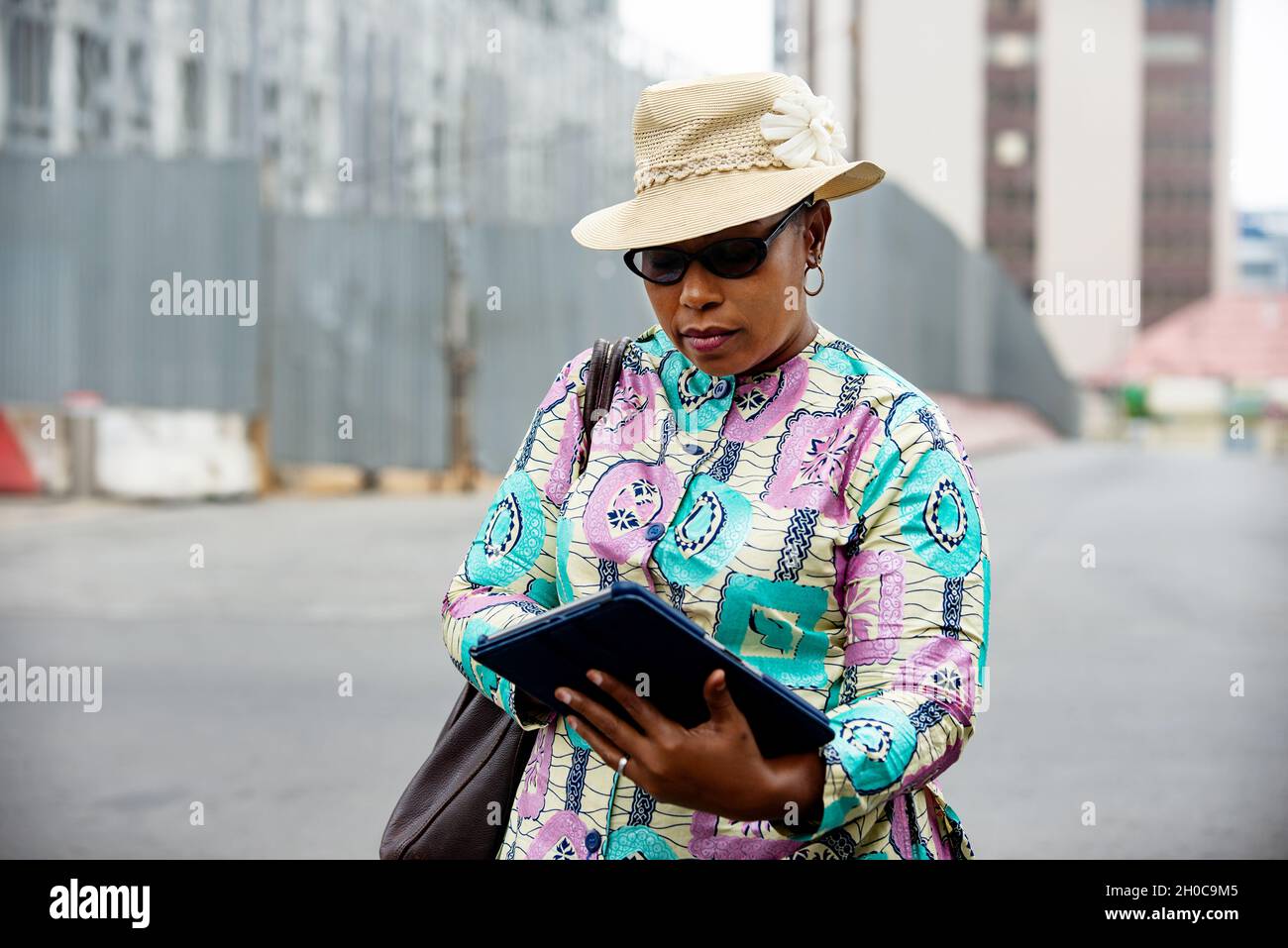 Elegante Afrikanerin, die auf der Straße in der Hand ihres Tablets steht, während sie sich ansieht, trägt einen schönen Lendenschurz und auf der Schulter eine Tasche Stockfoto