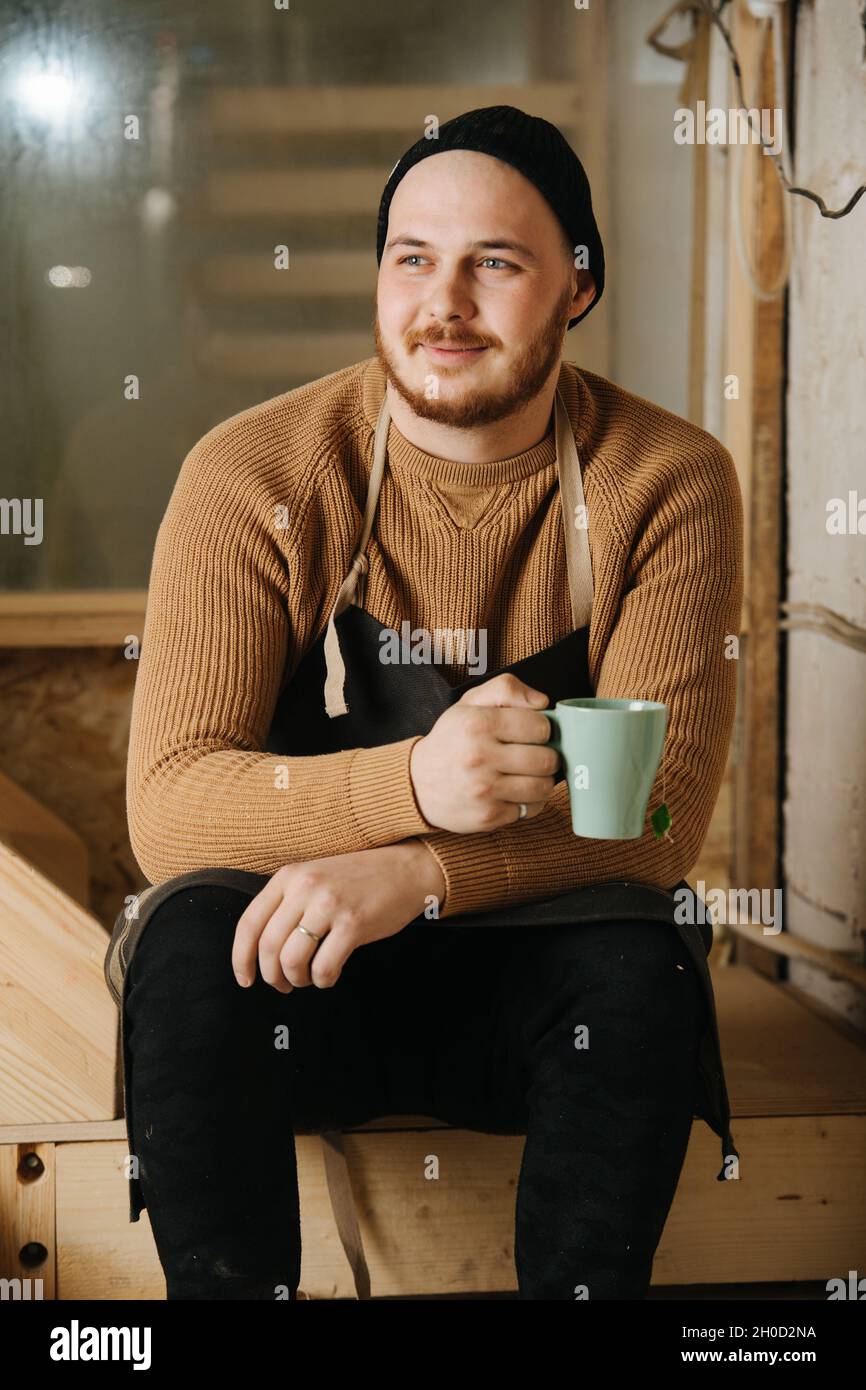 Porträt eines Zimmermanns in einer Uhrenmütze, der auf einer Holztreppe in einer Werkstatt sitzt und Tee aus dem Becher trinkt. Suchen Sie sich etwas anderes. Stockfoto