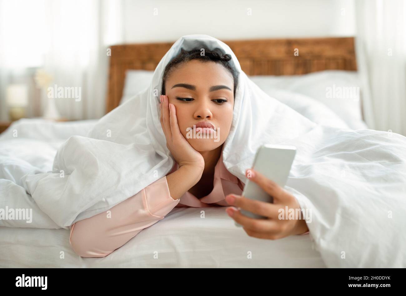 Verärgert junge schwarze Frau, die auf den Smartphone-Bildschirm schaut, während sie unter der Decke im Bett liegt und sich einsam oder deprimiert fühlt Stockfoto