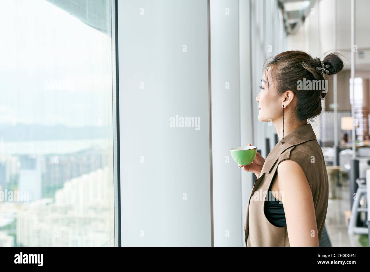 Reife asiatische Frau, die am Fenster steht und durch das Fenster schaut und eine Tasse Kaffee oder Tee hält Stockfoto