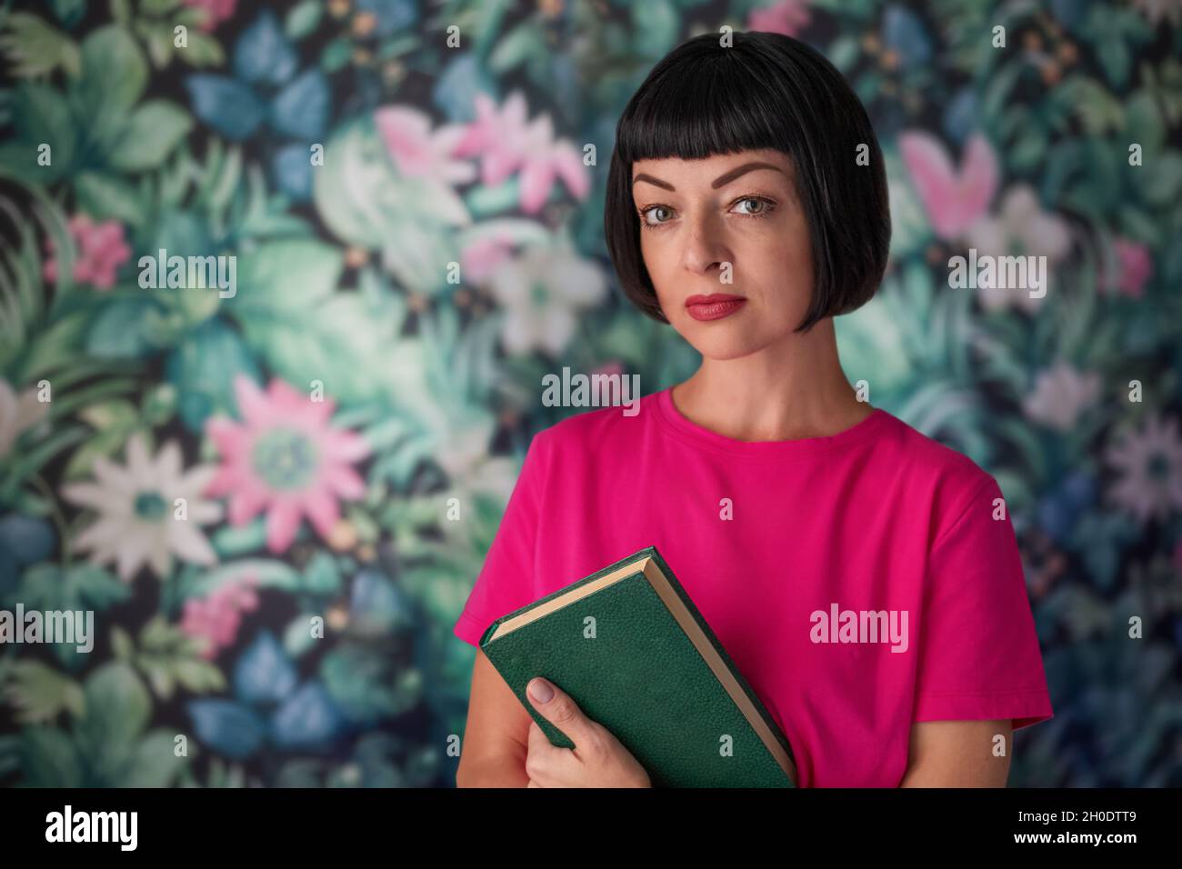 Eine Frau mittleren Alters hält ein Buch in ihren Händen. Steht vor dem Hintergrund einer mehrfarbigen Wand. Stockfoto