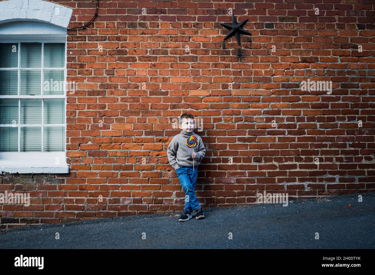 Kleiner Junge mit großem Lutscher, der sich an einer alten Backsteinmauer lehnt Stockfoto