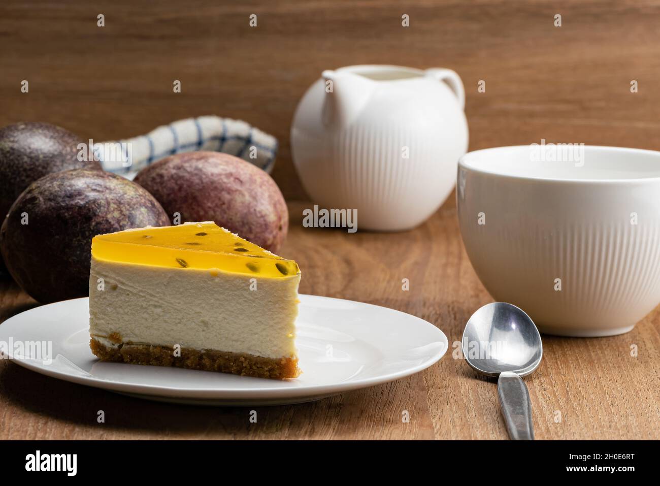 Blick auf köstlichen hausgemachten Passionsfrucht-Käsekuchen in weißer Keramikschale, eine Tasse Kaffee, reife Passionsfrucht, Metalllöffel und Milchtopf auf Holzknüppel Stockfoto