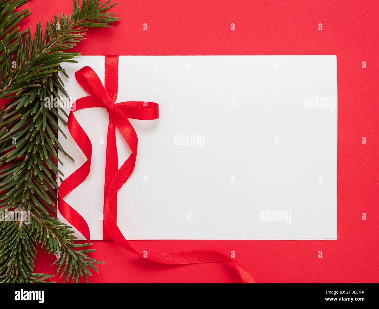 Weihnachtliche Geschenkanhänger weiß blank mit roter Schleife auf rotem Hintergrund. Papiernotiz und Tannengirlande, leerer Raum, Grußkartenvorlage. Neu Ja Stockfoto