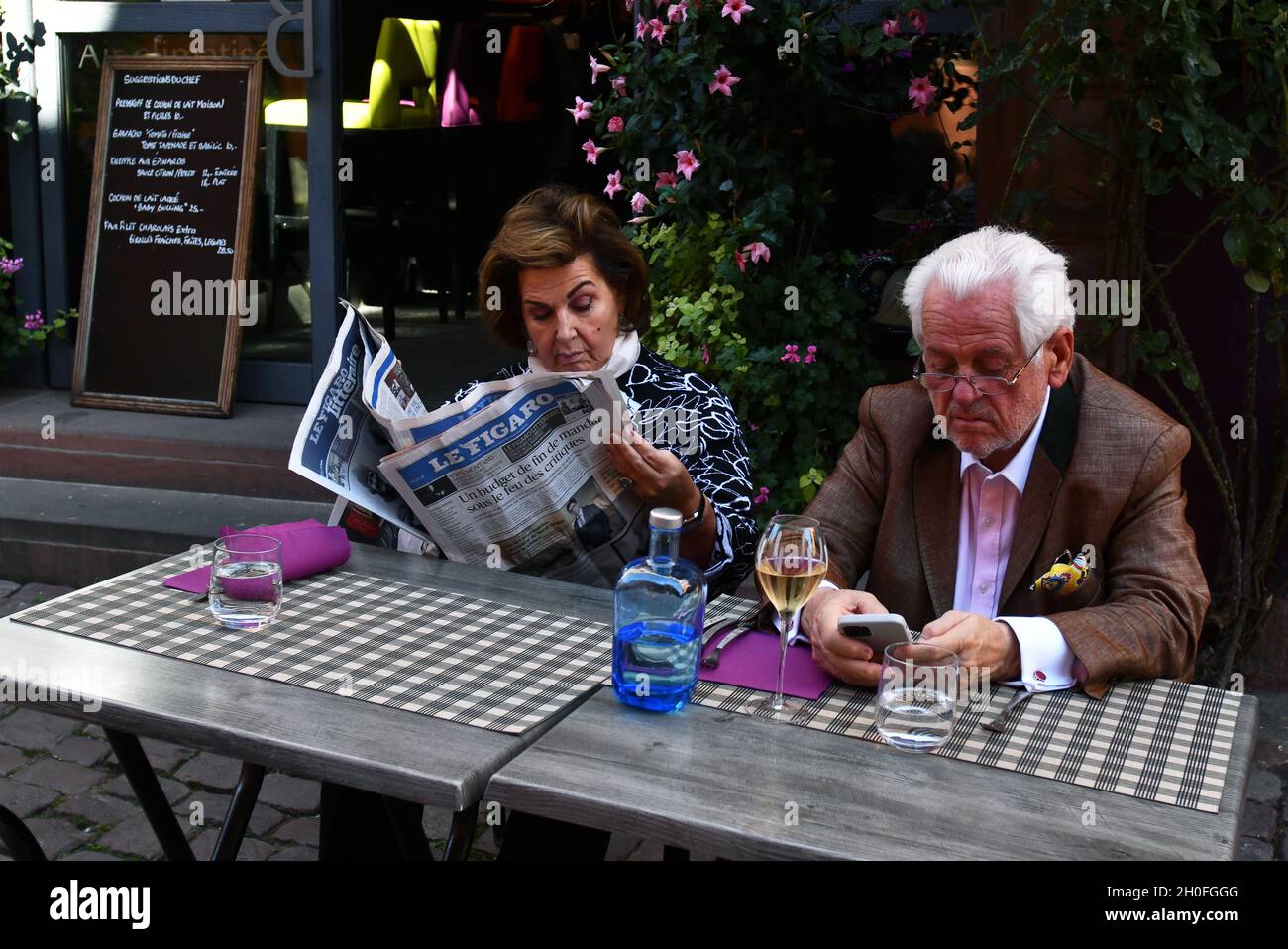 Eine Dame liest eine Zeitung, während der Mann sein Smartphone vor einem Café-Restaurant in Frankreich ansieht Stockfoto