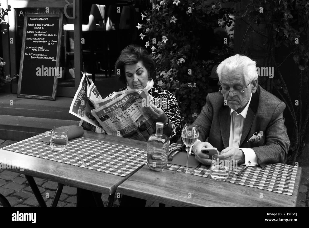 Eine Dame liest eine Zeitung, während der Mann sein Smartphone vor einem Café-Restaurant in Frankreich ansieht Stockfoto