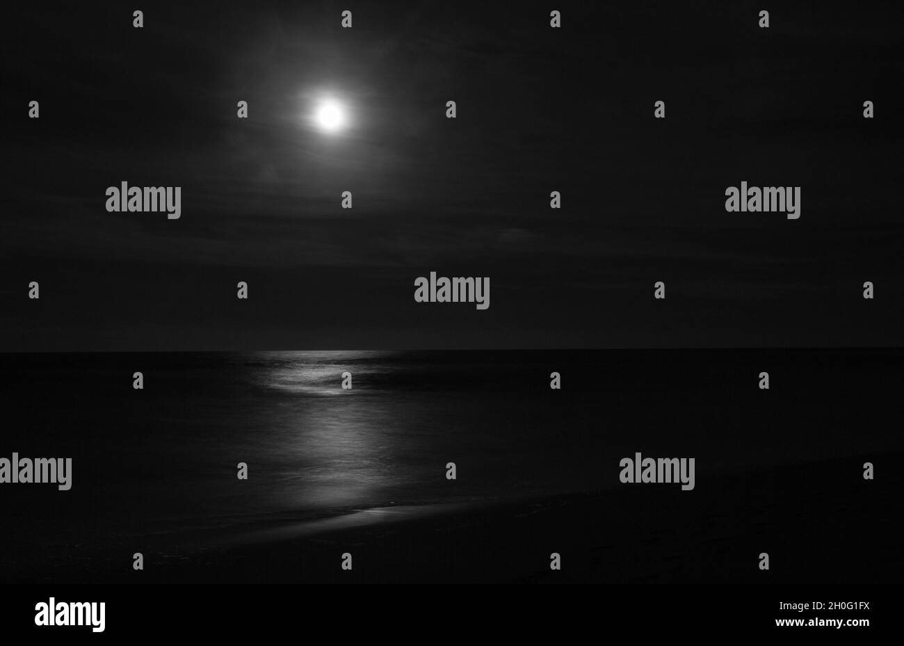 Nächtliche Seestücke und Brandung in Schwarz und Weiß mit hellem Mond, der auf der ruhigen Brandung und Küste scheint. Stockfoto