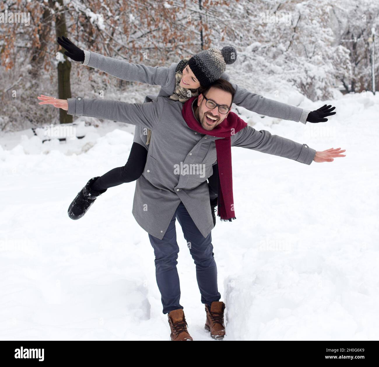 Glückliches lächelndes Paar genießt auf Schnee im Wald. Mann, der Mädchen auf dem Rücken vor weißen Bäumen hebt Stockfoto