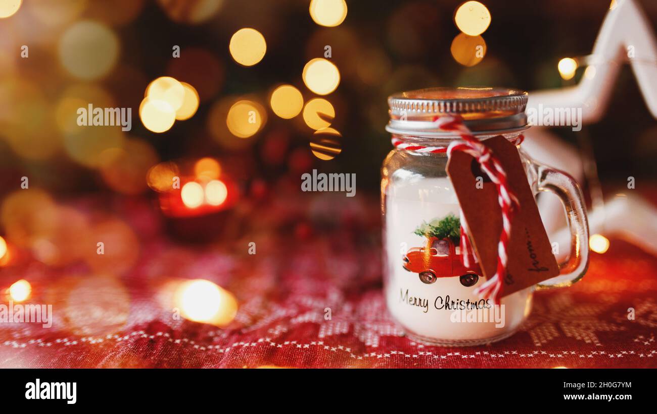 Festliche Weihnachtskerze in Form von Glas-Tasse oder Becher und anderen Dekorationen, verschwommener Hintergrund mit Weihnachtstanne Lichter, Girlanden und Kerzen. R Stockfoto