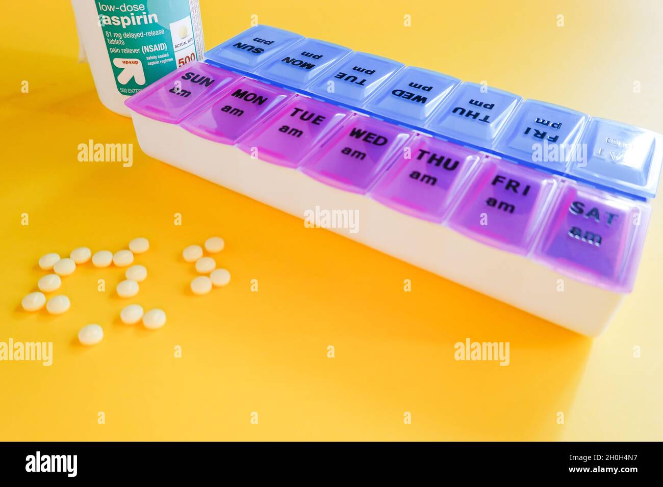 Bayer-Behälter mit 81-mg-Aspirin-Tablette mit niedriger Dosis und Sicherheitsbeschichtung auf gelbem Hintergrund mit täglicher Tablettenplanerbox Stockfoto