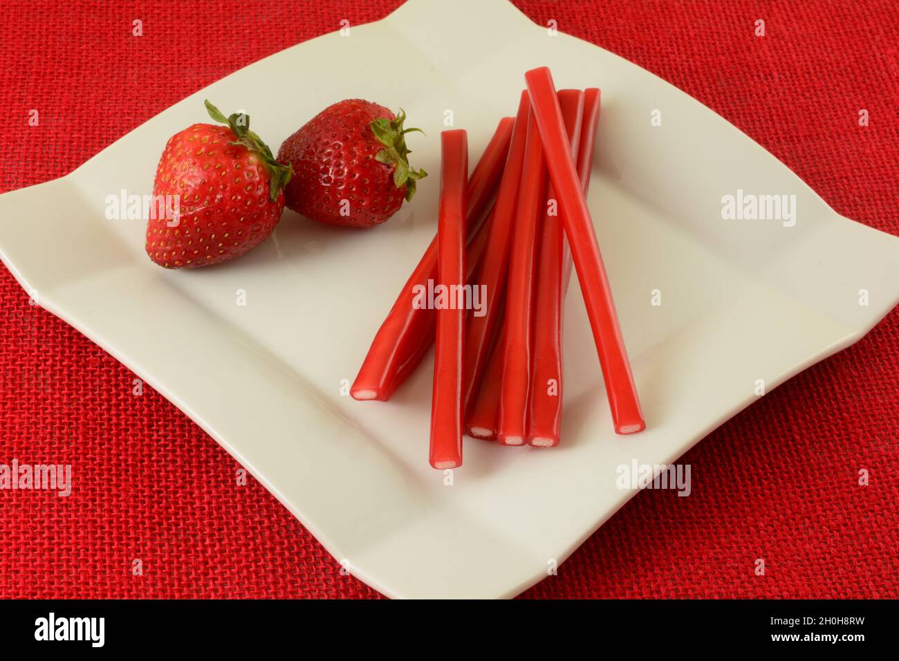 Erdbeer-Bleistift-Stick Gummy Candy mit frischen, rohen Erdbeeren auf weißem Dessertteller auf roter Tischdecke Stockfoto