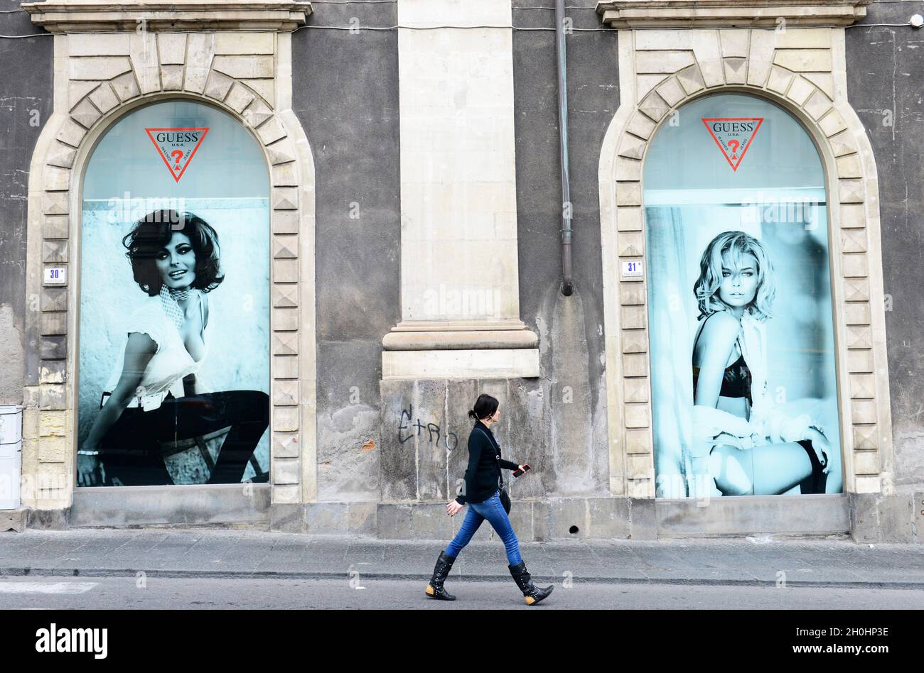 Eine Italienerin, die unter einer großen Werbung für das Bekleidungsgeschäft Guess im Palazzo Tezzano in Catania, Italien, läuft. Stockfoto