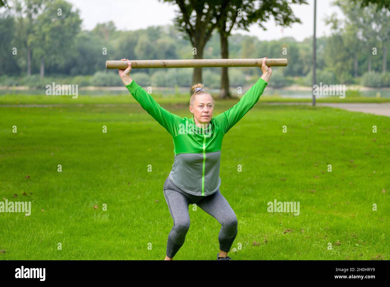 Fit muskulösen Frau mit einem Balken aus Holz in einem üppigen grünen Frühlingsfeld heben es über ihren Kopf in einer Hocke Position, um ihren Kern mu zu stärken Stockfoto