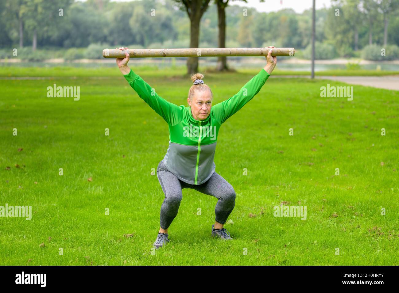 Fit muskulösen Frau mit einem Balken aus Holz in einem üppigen grünen Frühlingsfeld heben es über ihren Kopf in einer Hocke Position, um ihren Kern mu zu stärken Stockfoto