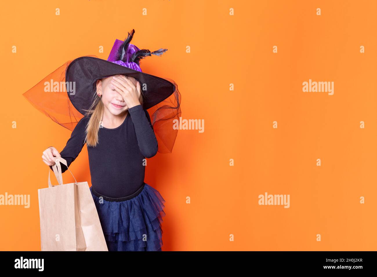 Ein wunderschönes Mädchen in einem schwarzen Kleid und einem Hexenhut hält eine Bastelpapiertasche, die ihr Gesicht mit der Hand bedeckt und sich wundert, auf einem orangefarbenen Hintergrund. Die CO Stockfoto