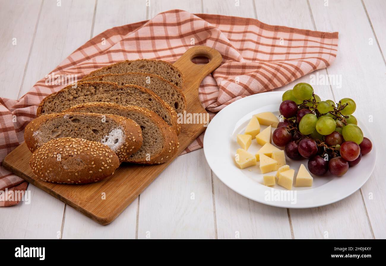 Seitenansicht von geschnittenem braunem Saatkäfelchen auf Schneidebrett auf kariertem Tuch und einem Teller mit Käse und Trauben auf Holzhintergrund Stockfoto