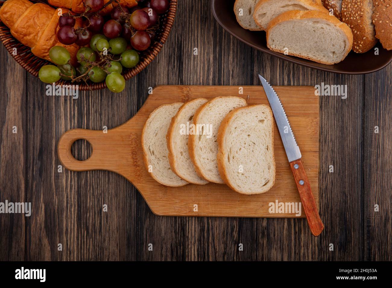 Draufsicht auf Brote, wie in Scheiben geschnittene braune Kob und weiße in einer Schüssel und auf einem Schneidebrett mit Messer und einem Korb mit Croissant-Trauben auf einem Holzbackgroun Stockfoto