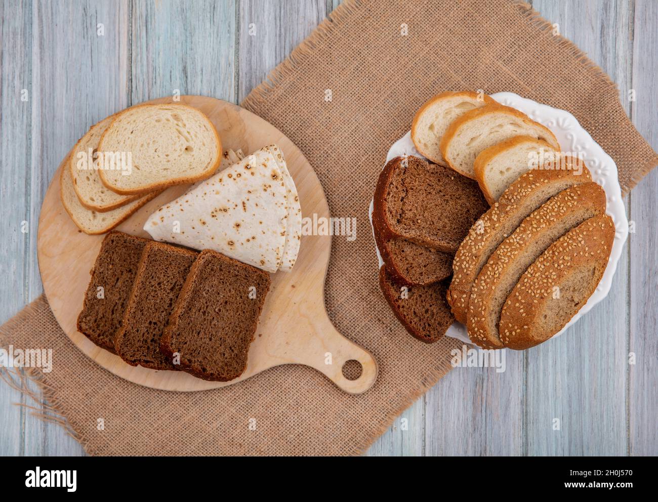 Draufsicht auf Brote, wie in Scheiben geschnittenes Roggenbrot mit weißem Saatkorn und Fladenbrot auf Schneidebrett und in Teller auf Sacktuch auf Holzhintergrund Stockfoto