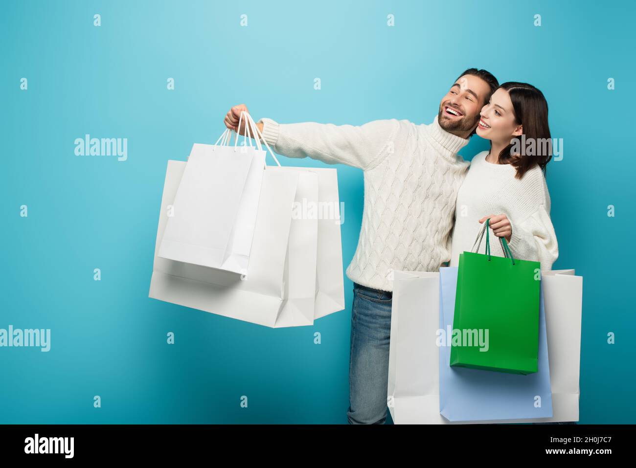 Lächelndes Paar in weißen Pullovern, das wegschaut, während es Einkäufe auf Blau hält Stockfoto