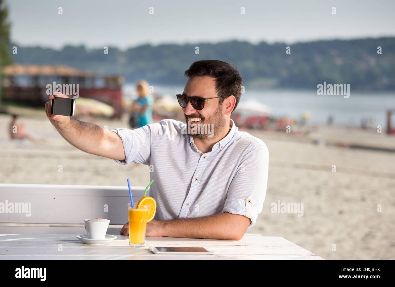 Ein hübscher Mann mit Sonnenbrille, der am Strand ein Handy zum Webchatten benutzt. Genießen Sie am Fluss Stockfoto