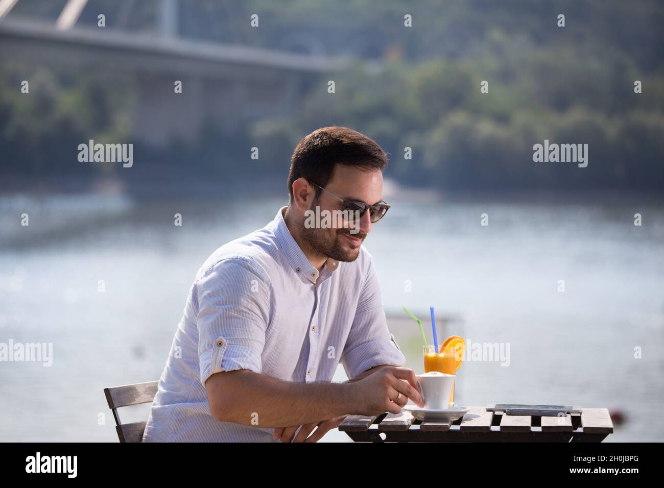 Schöner Mann, der morgens Kaffee und Orangensaft im Café am Flussstrand trinkt Stockfoto